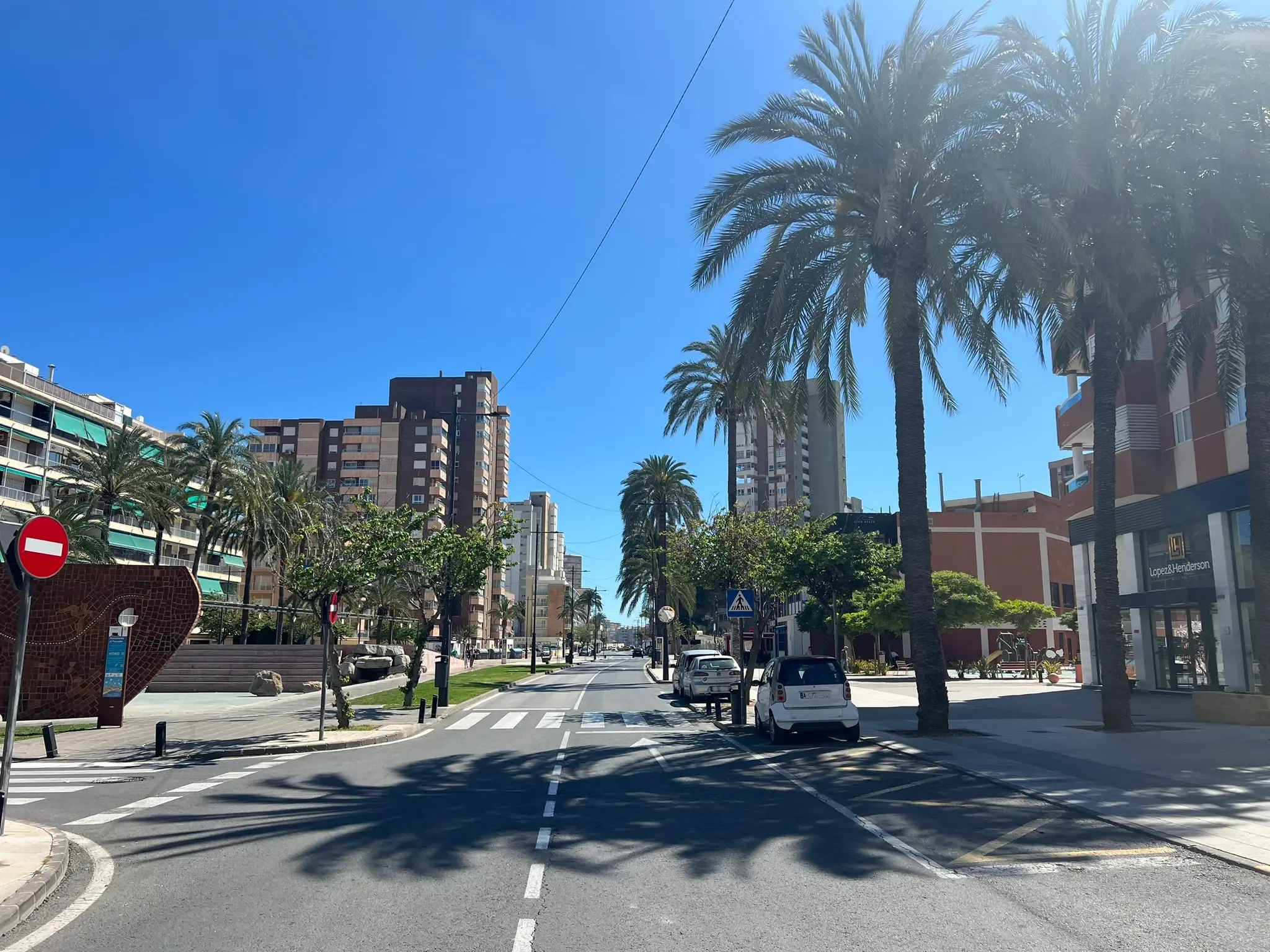 A sun-drenched wide street in El Campello leading towards the coast. Tall palm trees line the sidewalk, casting long shadows across the asphalt. Modern multi-story apartment buildings and local businesses stand under a clear, bright blue sky.